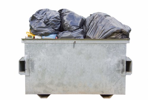 A large metal skip bin filled with tightly packed black rubbish bags, situated outdoors on a paved surface. The skip appears to be made of galvanized steel with a slightly textured finish and has two recessed side handles. Several bags are stacked above the rim, some leaning against each other, with their glossy, crinkled surfaces visibly reflecting light. The bins' surroundings are not fully visible, but the setting suggests an industrial or commercial area, possibly in Southwark, where rubbish removal services like those provided by Office Clearance Southwark operate. The overall scene indicates a collection of waste awaiting disposal or collection, consistent with professional rubbish clearance activities in London and the surrounding boroughs.