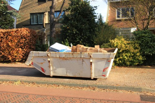 A large white metal skip filled with various types of cardboard boxes and packaging material, positioned on a paved residential street in Southwark, London. The skip is situated on the pavement, with some rust visible along its edges and sides. In the background, there are residential houses with brick facades, pitched roofs, and multiple windows, surrounded by trees and shrubs, indicating a suburban neighbourhood. The scene is captured during daylight, with natural sunlight casting shadows on the street and skip, highlighting the textured surfaces of the cardboard and the skip's weathered metal finish. This image relates to rubbish removal services offered by Office Clearance Southwark, demonstrating typical waste disposal activities in the local area.